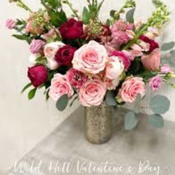 Pink and red rose arrangement with accent blooms in a tall metallic vase on a light surface.