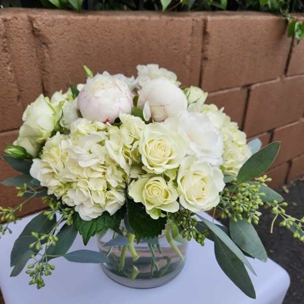 Low round arrangement of white roses, hydrangeas, and peonies in a clear glass vase