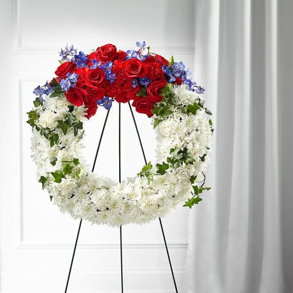 Circular funeral wreath of red, white, and blue flowers on a black stand