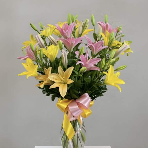 Tall clear vase of pink and yellow lilies with buds, tied with a pink and yellow ribbon bow.
