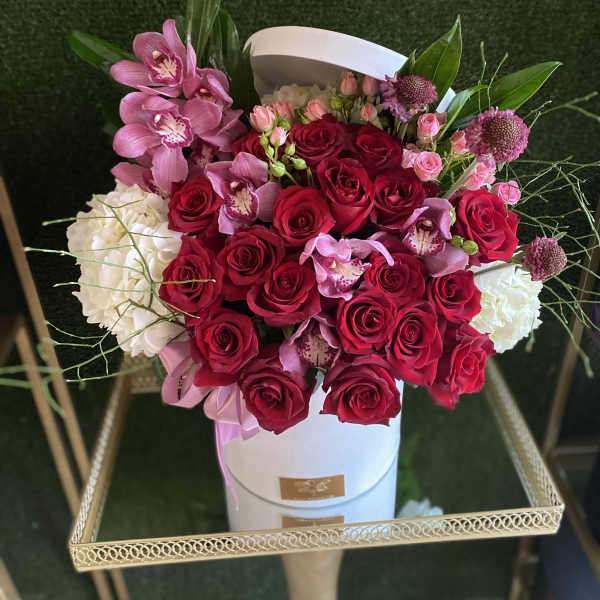 Round white hat box filled with red roses, pink orchids, and white blooms on a gold stand