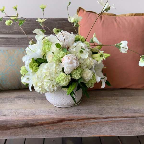 White floral arrangement in a white vase with pink and blue pillows behind it