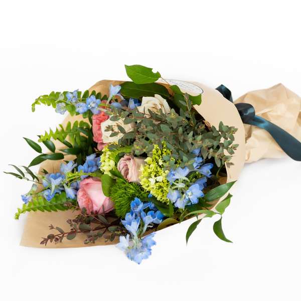 Wrapped bouquet of pink and cream roses with blue accent flowers in kraft paper tied with a dark ribbon