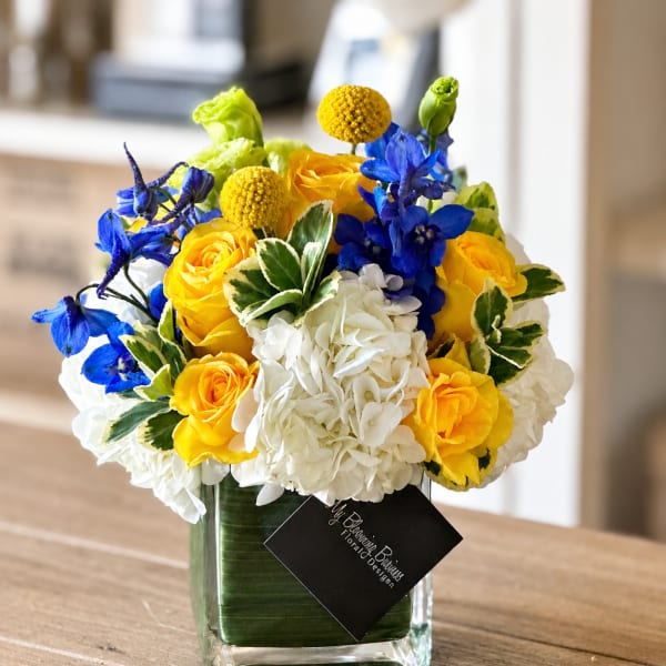 Yellow roses, white hydrangeas, and blue flowers in a square glass vase