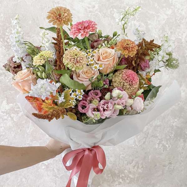 Handheld bouquet of peach, pink, and white flowers wrapped in white paper with a pink ribbon.
