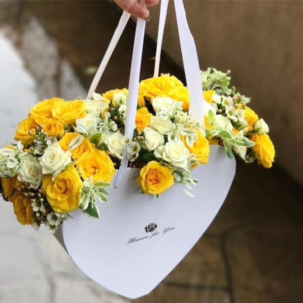 Yellow and white rose bouquet in a white heart-shaped box with ribbon handles