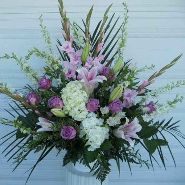 Tall floral arrangement with pink lilies, purple roses, and white hydrangeas in a white vase.
