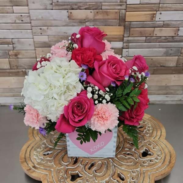Compact arrangement of hot pink roses, white hydrangea, and pink carnations in a heart-print cube container.