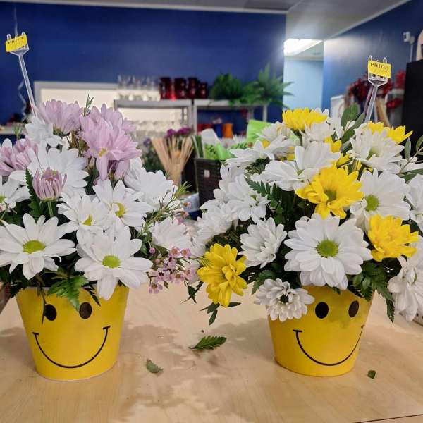 Two daisy arrangements in yellow smiley-face pots, one with pink and white blooms and one with white and yellow blooms.