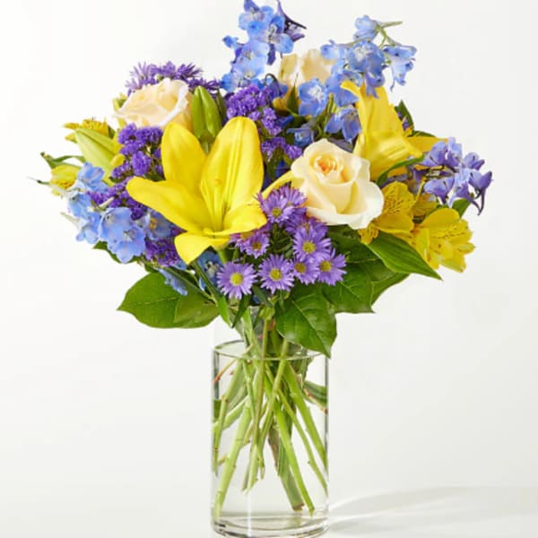 Bouquet of yellow lilies, cream roses, and purple flowers in a clear glass vase