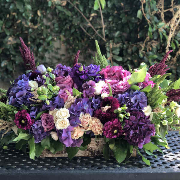 Low floral arrangement of purple, pink, and white blooms in a wooden box