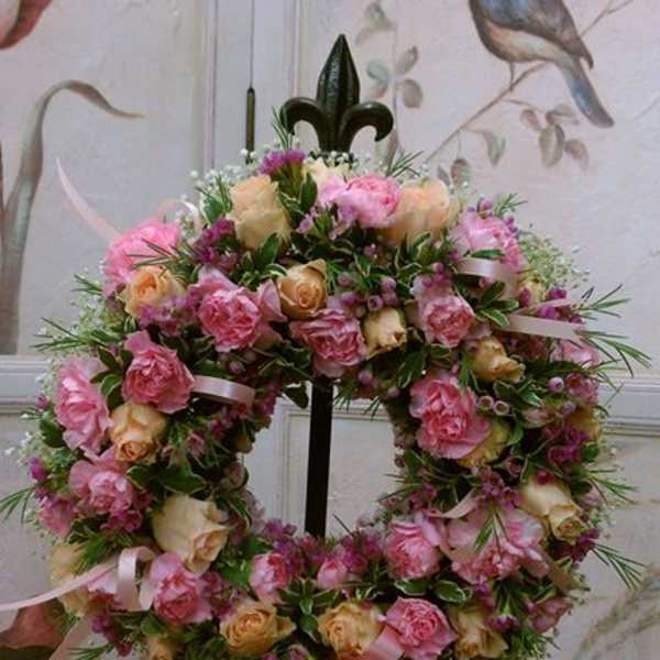 Round wreath of pink and peach roses with small purple blossoms and pink ribbons on a stand