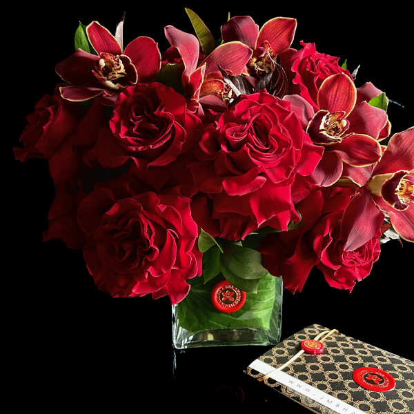 Red roses and orchids arranged in a clear square vase beside a patterned gift box