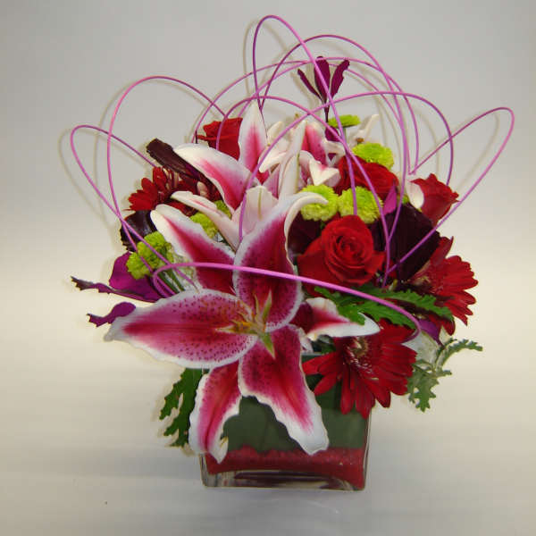 Pink lilies and red roses in a square glass vase with looping purple accents