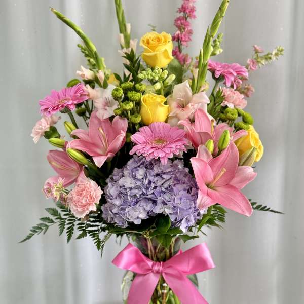 Mixed bouquet of pink lilies, yellow roses, and lavender hydrangea in a glass vase with a pink ribbon