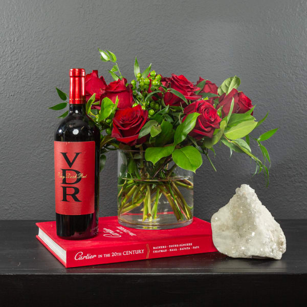 Red roses in a glass vase beside a wine bottle and a red book