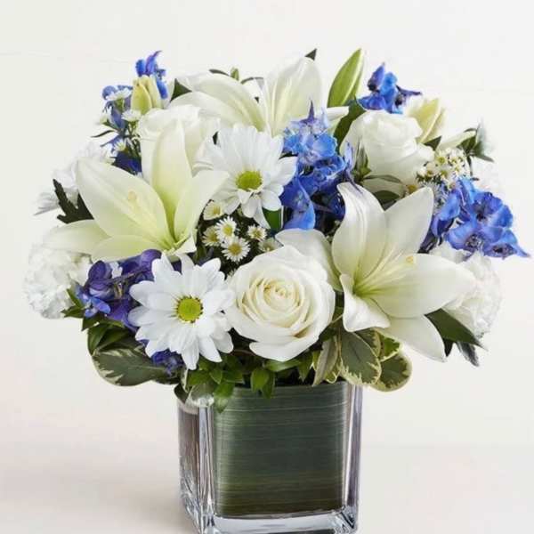 White lilies, roses, and daisies with blue flowers in a square glass vase