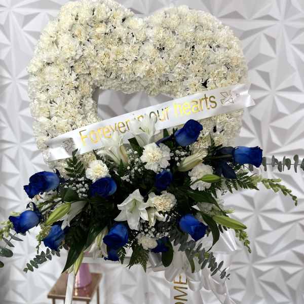 White floral heart wreath with blue roses and lilies, plus a ribbon reading "Forever in our hearts"