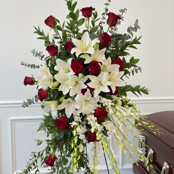Standing funeral spray with red roses, white lilies, and trailing orchids beside a casket