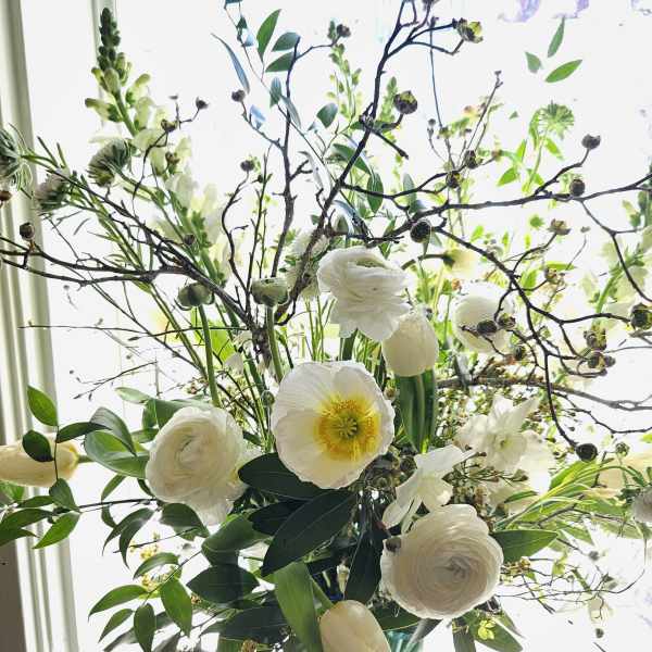 Tall arrangement of white flowers and twiggy branches in a blue glass vase by a bright window