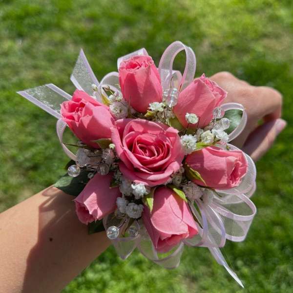 Pink rose wrist corsage with white filler flowers, ribbon loops, and bead accents on a person's wrist