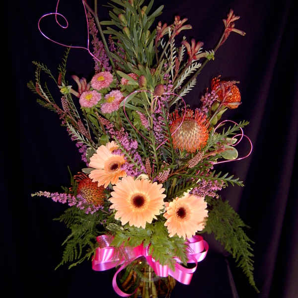 Tall mixed bouquet with peach gerberas, pink blooms, and a pink ribbon in a glass vase