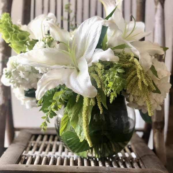 White lilies and mixed white and green flowers in a round glass vase.