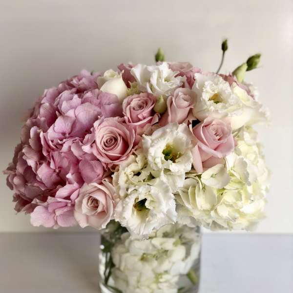 Low round arrangement of pink hydrangeas, pale pink roses, and white flowers in a clear glass vase.