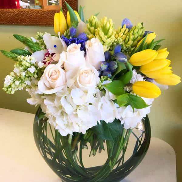 Bouquet of white roses, yellow tulips, and blue flowers in a round glass vase