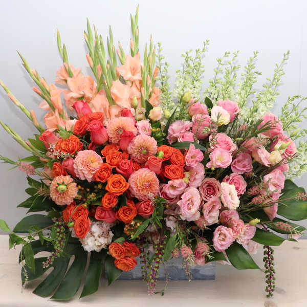 Large rectangular arrangement of peach gladiolus, orange dahlias, and pink roses in a low wooden container