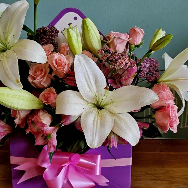 Pink roses and white lilies arranged in a purple gift box with a pink ribbon