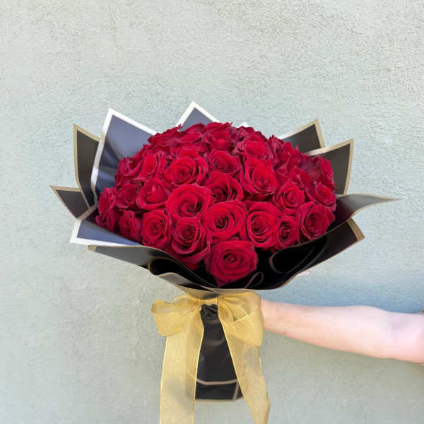 Arm holding a large bouquet of red roses wrapped in black paper with a gold bow.