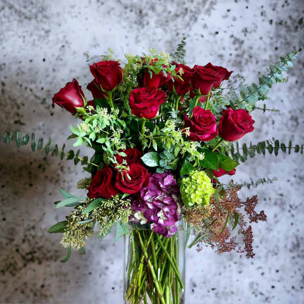 Red roses arranged in a clear glass vase with mixed greenery and purple hydrangea