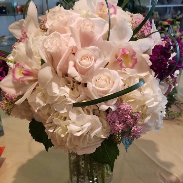 Round bouquet of pale pink roses, white hydrangeas, and orchids in a clear glass vase