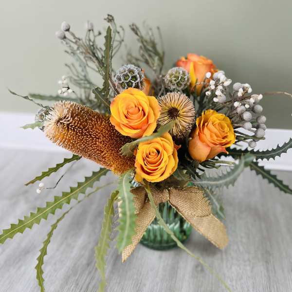 Orange roses in a glass vase with dried seed pods and a burlap ribbon