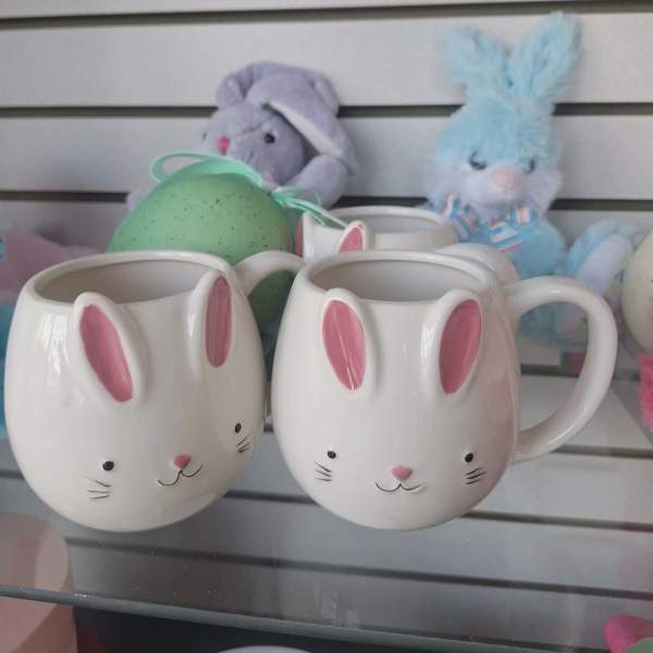 Two white bunny-face mugs with pink ears on a glass shelf with pastel bunny toys behind.