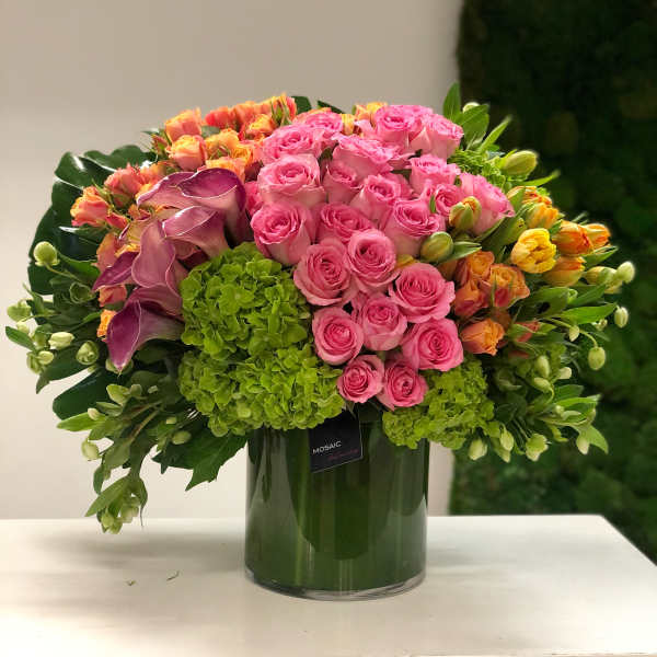 Lush arrangement of pink roses, green hydrangeas, orange tulips and purple calla lilies in a glass cylinder vase