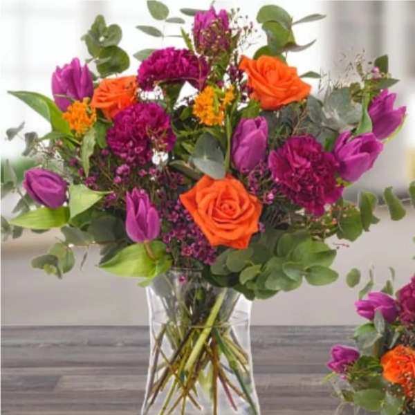 Bouquet of orange roses, purple tulips, and magenta flowers in a clear glass vase