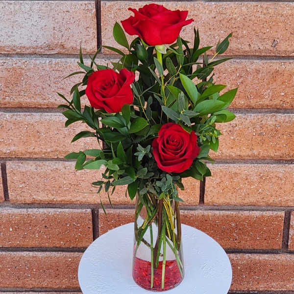Three red roses in a clear glass vase with greenery