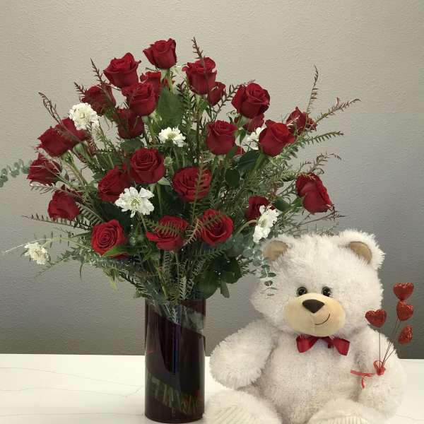 Red roses in a tall vase beside a white teddy bear holding heart picks