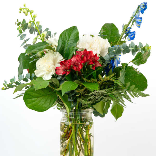 Red, white, and blue mixed flower arrangement with lush foliage in a clear glass jar vase