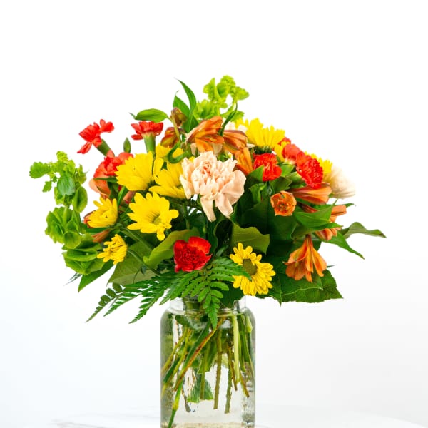 Bright mixed bouquet of yellow daisies, orange alstroemeria, and red and peach carnations in a clear glass jar.