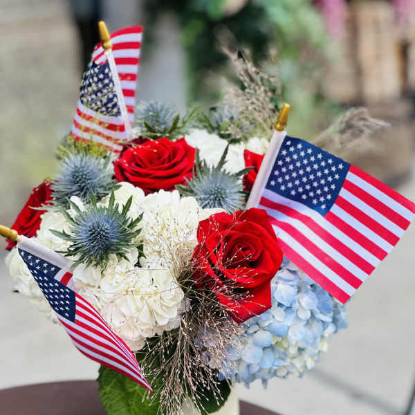 Red roses and blue hydrangeas in a white vase with small American flags
