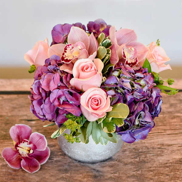 Low round arrangement of pink roses, cymbidium orchids, and purple hydrangeas in a silver vase.