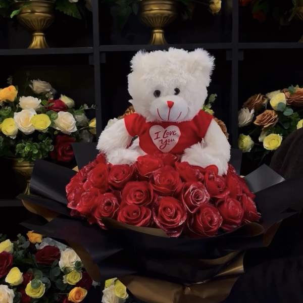 White teddy bear in a red shirt sitting on a bouquet of red roses