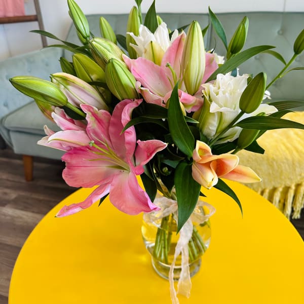 Bouquet of pink, white, and peach lilies with buds in a clear glass vase on a yellow table