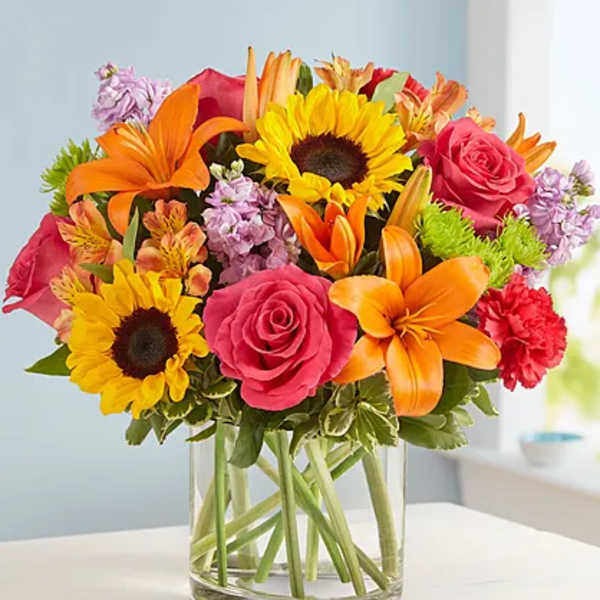 Bright mixed bouquet in a clear glass vase with sunflowers, roses, lilies, and carnations