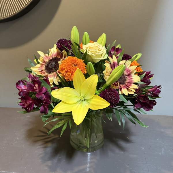 Mixed bouquet with yellow lilies, roses, sunflowers, and purple blooms in a glass vase