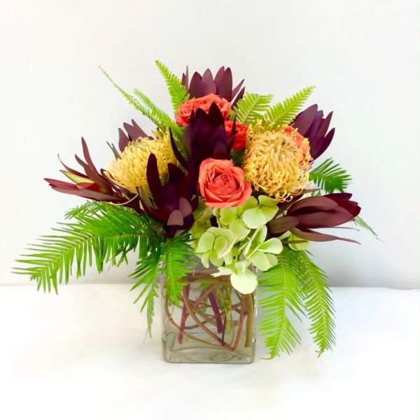 Tropical bouquet with coral roses and burgundy blooms in a square glass vase