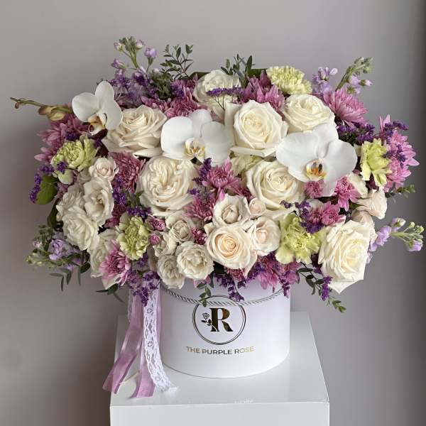 Round hat box of ivory roses, white orchids, and pink and purple flowers with trailing ribbons.